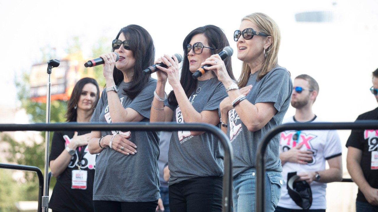 Three women wearing gray T-shirts each hold microphones in their left hands.
