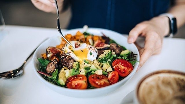 A person holds a fork over a white bowl with a salad topped with tomato, avocado, sweet potato, mushroom, fig and egg.