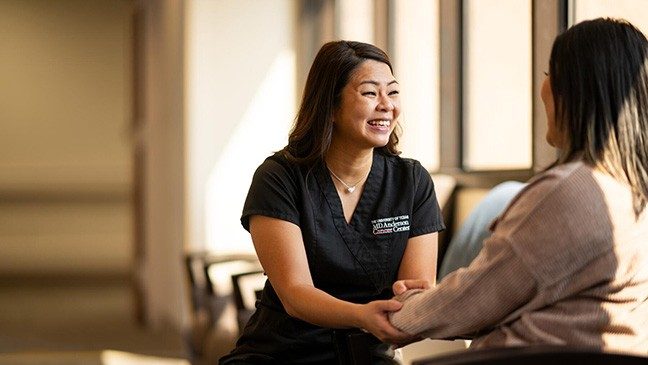 A woman in black scrubs smiles and holds the hand of the woman she is speaking with.