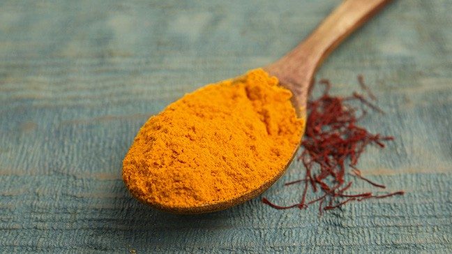 A wooden spoon holding orange saffron powder. Red saffron threads sit beside the spoon on a blue wooden surface.