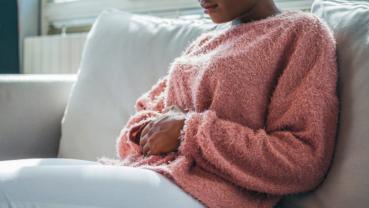 Black woman in a fuzzy pink sweater and white fitted pants holding her stomach in pain on a white couch