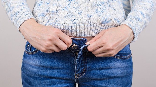 Waist-level photo of slender white sweater-wearing woman's hands struggling to button up her jeans