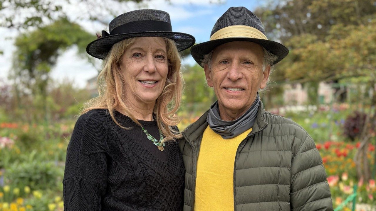 A woman and man wearing hats stand in a garden of colorful flowers