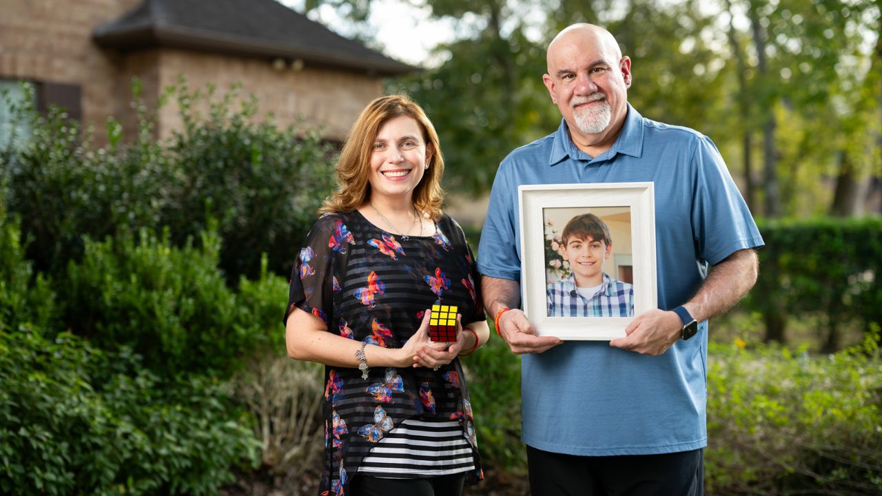 A woman and man stand together. The man holds a photo of a young boy. The woman holds a Rubik's cube.