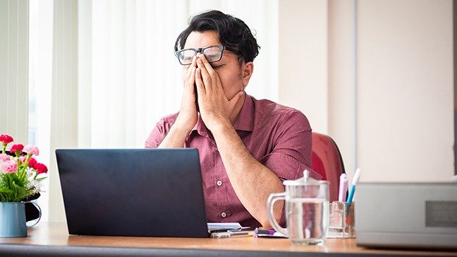 Young adult man in front of laptop places both hands on his face in exasperation 