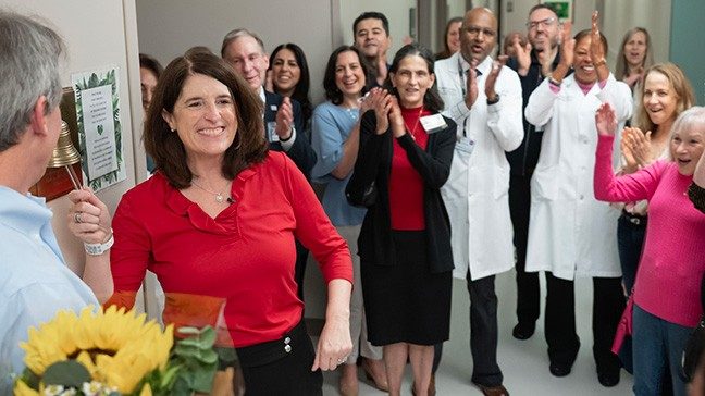 Woman in red shirt smiles as she rings a bell surrounded by a crowd of people clapping.