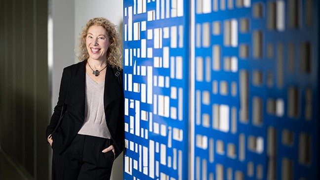 Woman with curly blonde hair wearing a black blazer and pants smiles as she leans against a wall with blue geometric artwork.