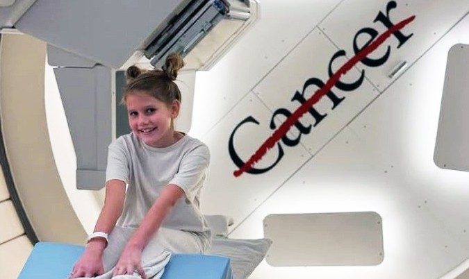 Young girl smiles while sitting near gantry in between proton therapy treatments