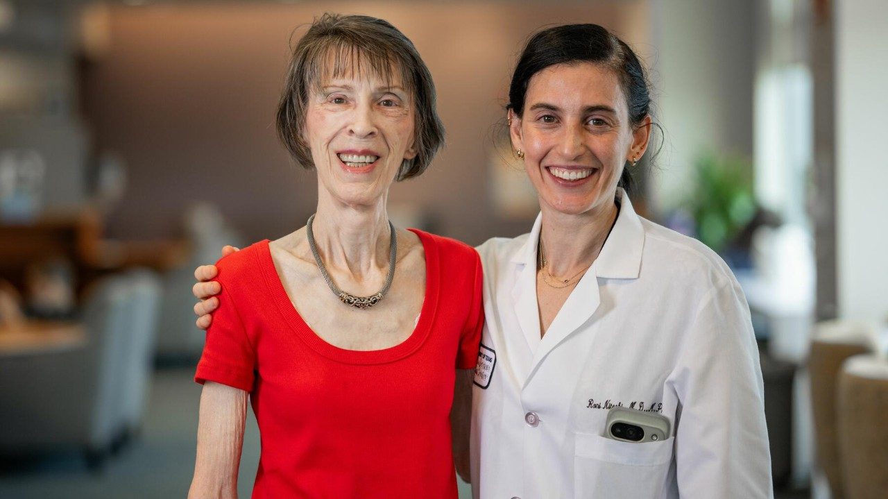 Woman wearing a bright red shirt stands next to a female doctor in a lab coat