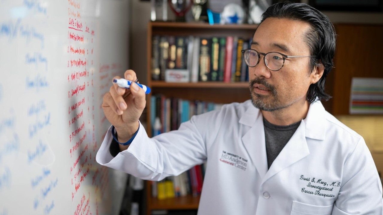 A man in a white lab coat writes with a marker on a dry erase board.