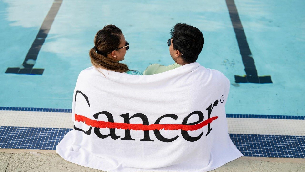 A man and a woman sit next to a pool wrapped in a beach towel that has the word cancer with a red strikethrough 