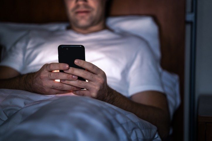 A man lies in bad, illuminated by the glow of a cell phone, How to cope with insomnia during cancer treatment