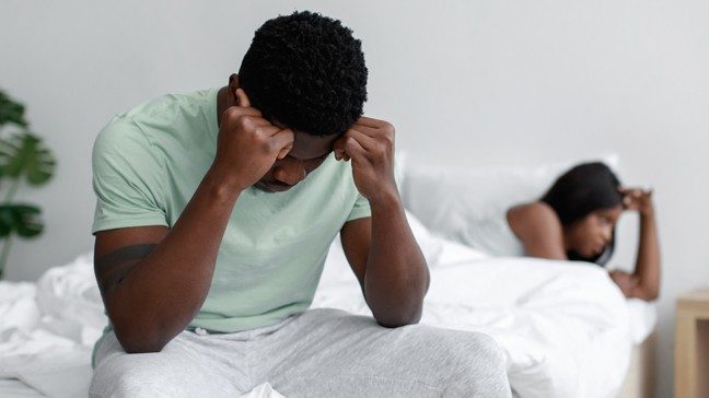 Visibly frustrated unidentified man sits on edge of bed with blurry woman in background