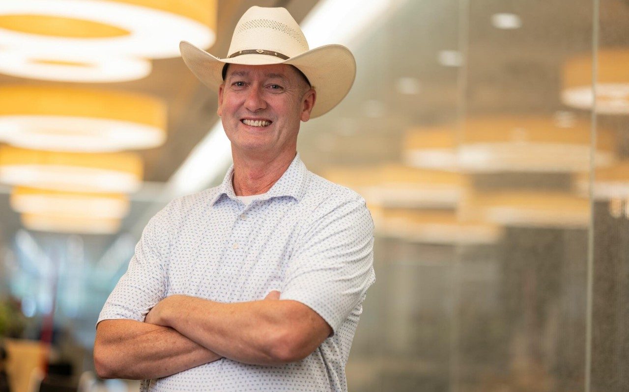 Man wearing a cowboy hat smiles while standing with his arms crossed 