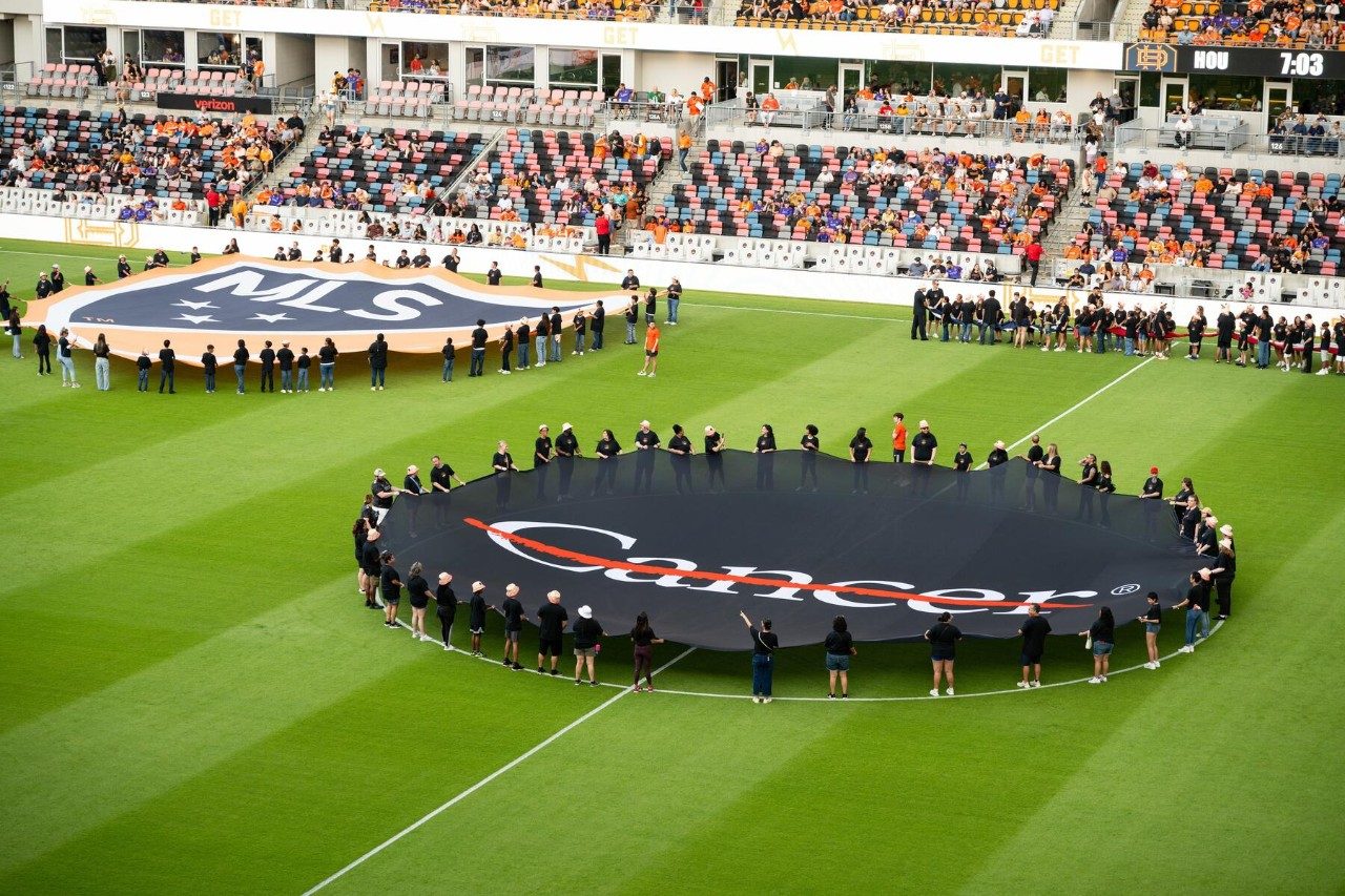Cancer survivors hold up a giant center circle banner showing the MD Anderson cancer strikethrough at Shell Energy Stadium