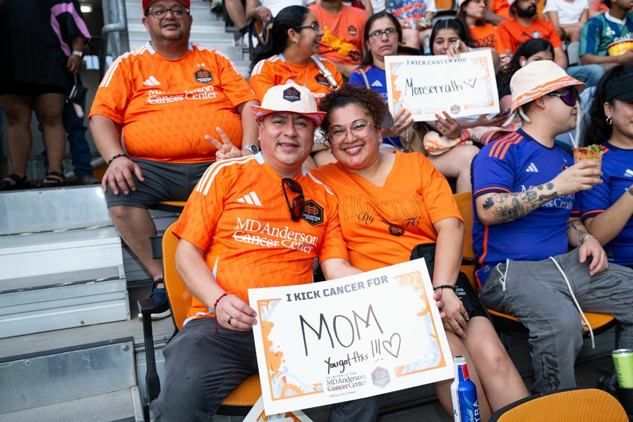 Two fans hold saying saying I kick cancer for Mom while sitting in the stands at Houston Dynamo FC Kick Cancer Night