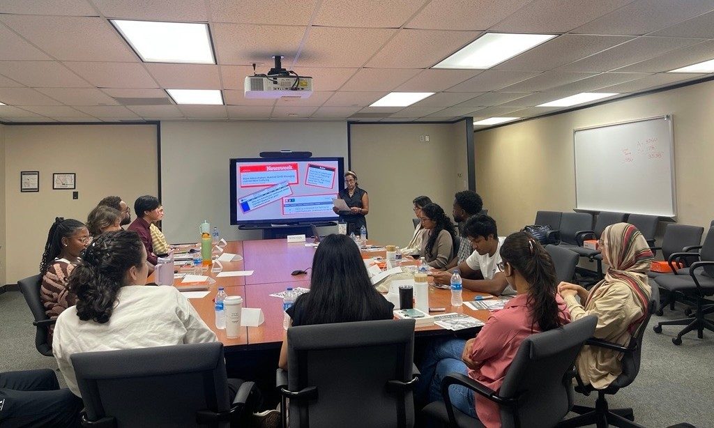 a group of students sitting at a table while someone presents at the front of a room