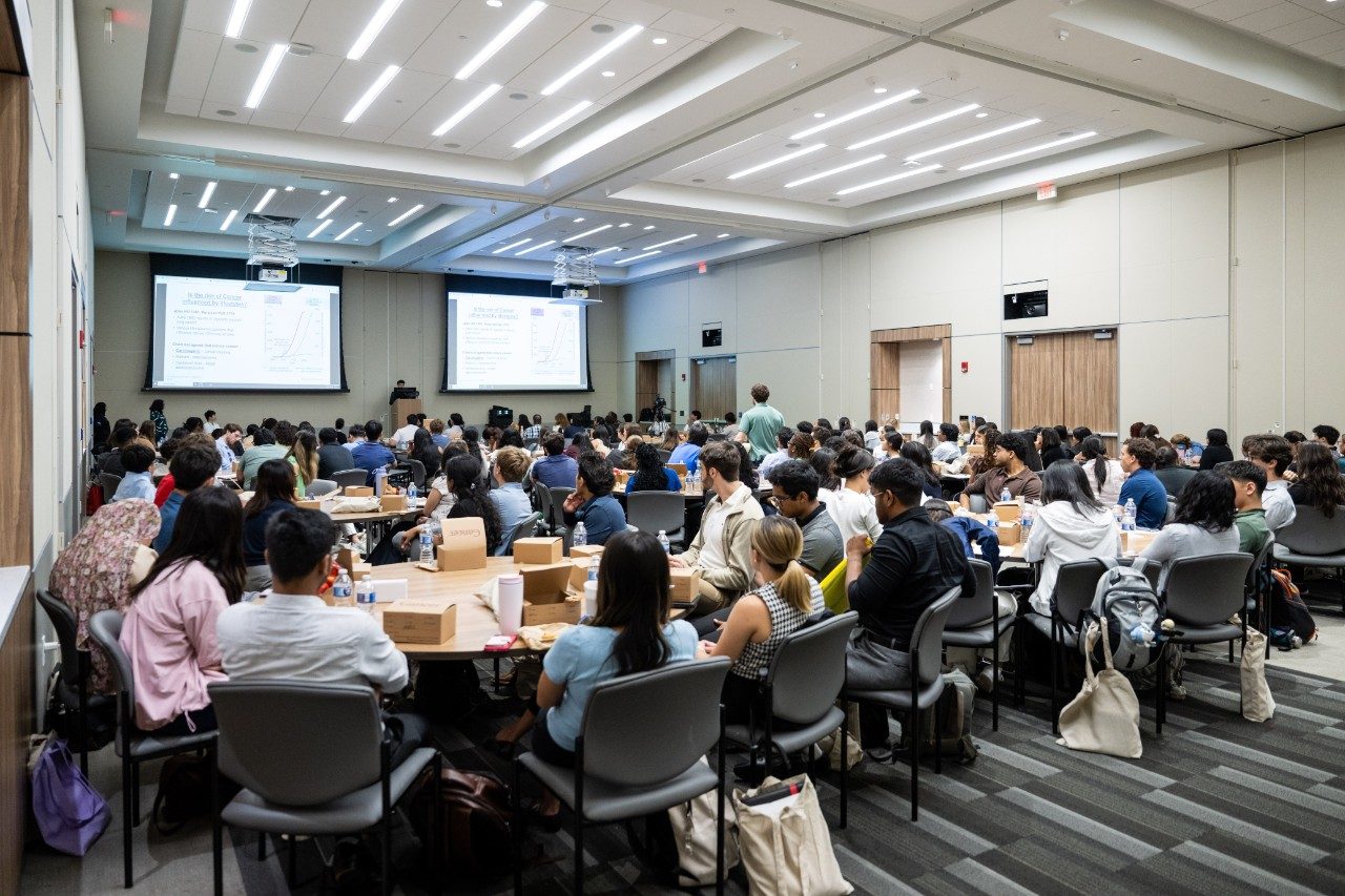 A room full of CATALYST Summer Training Program students sitting at round tables watching a lecture at the front of the room with projection screens to the left and right of the podium