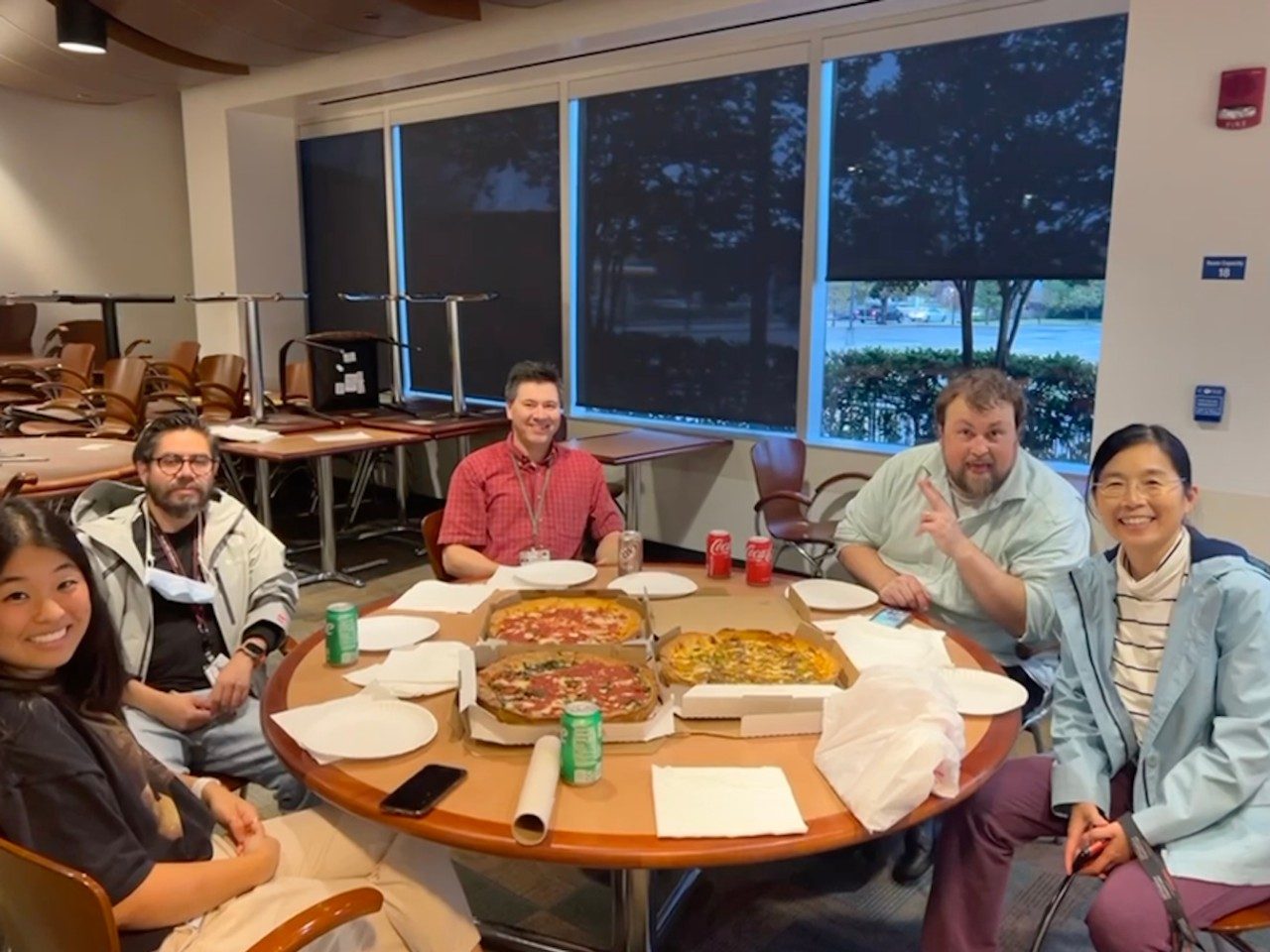 Five people sitting around a round table with three boxes of pizza on the table
