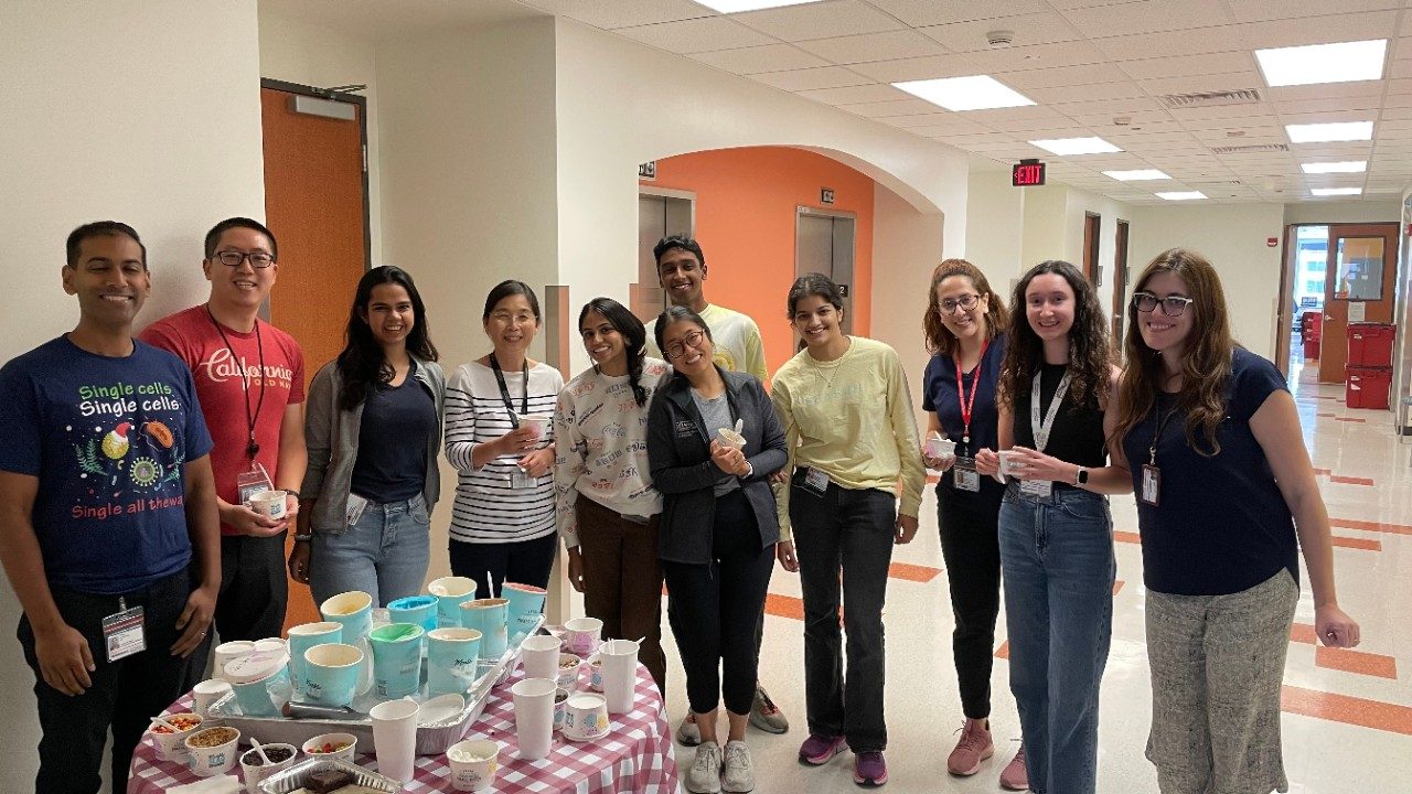Eleven people standing in a row in a hallway with a variety of ice cream and toppings on a table in front of them