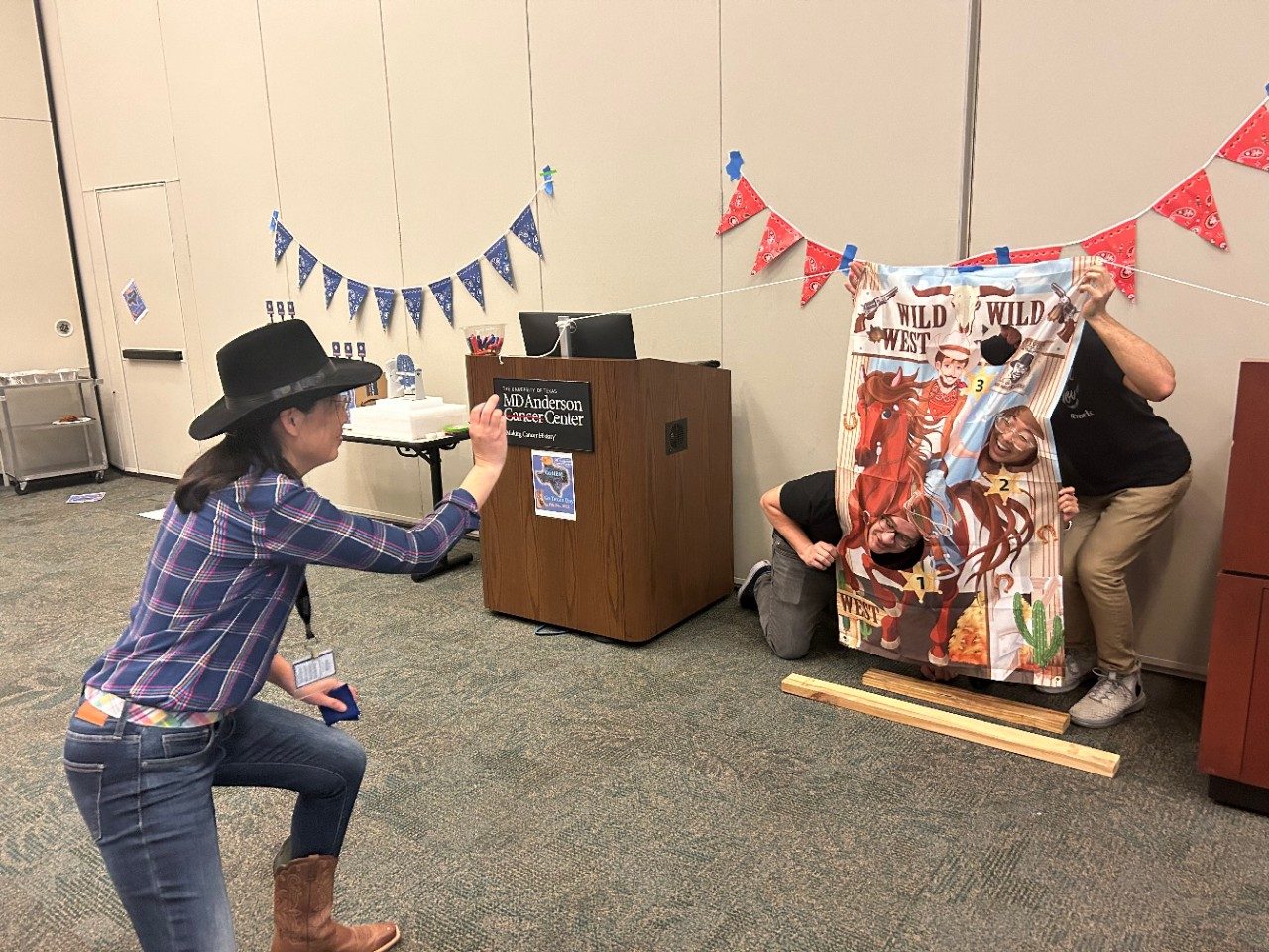 A person dressed up in a cowboy hat and plaid shirt waves towards a group of people standing behind a Wild, Wild West sign