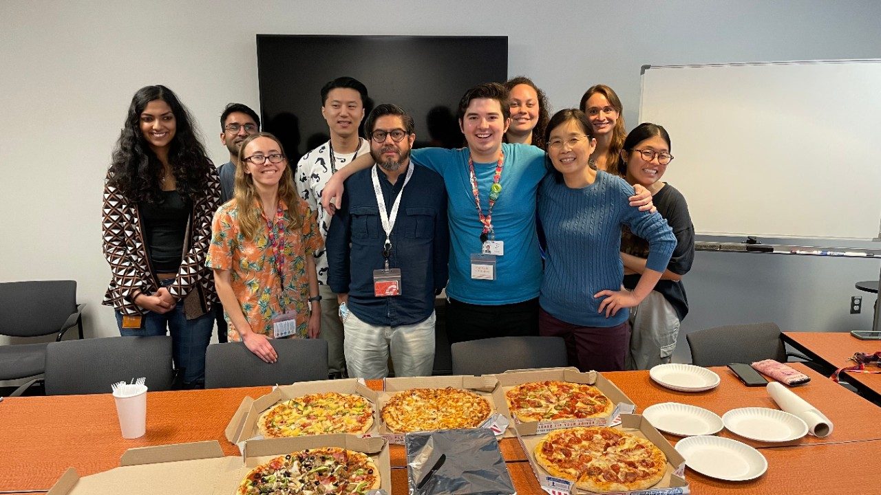 A group of people pose behind a rectangular conference room table with five pizzas on it