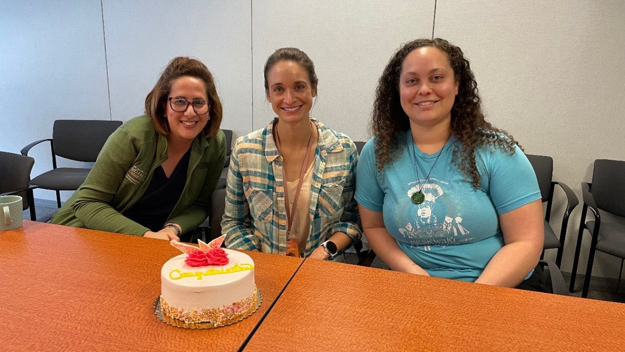Three women seated at a conference room table with a cake that reads "Congratulations" on the table in front of them