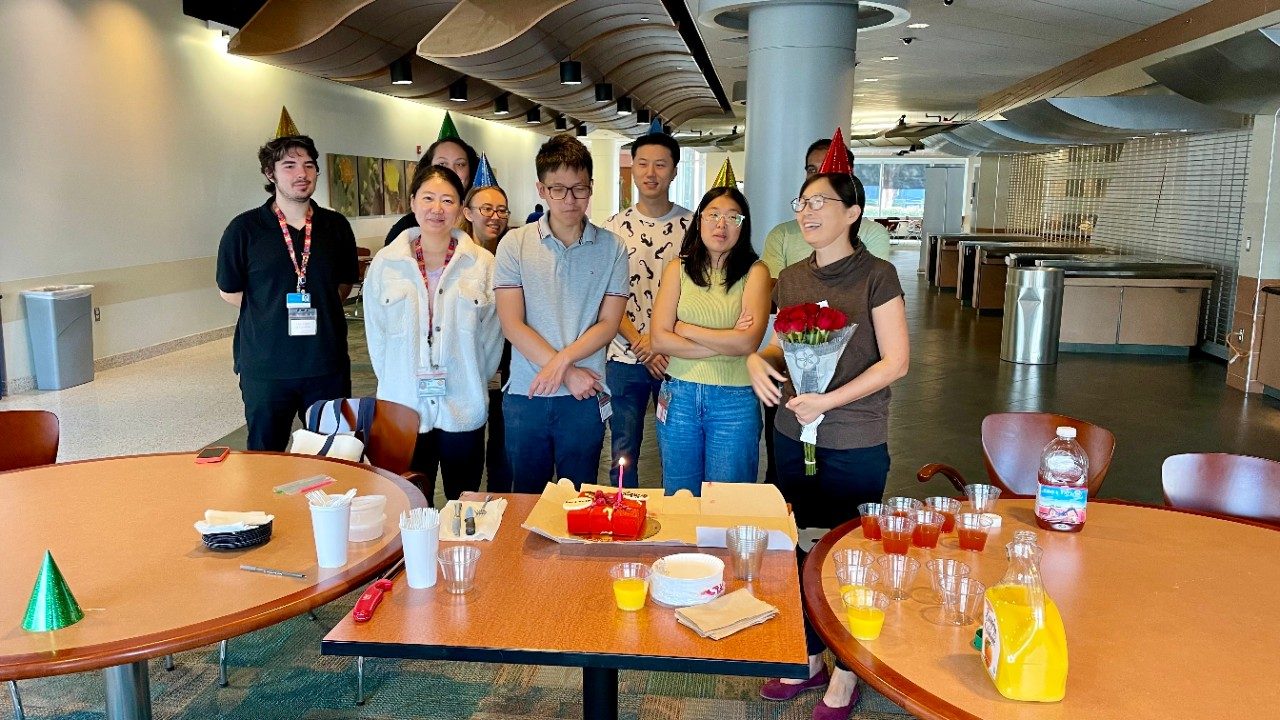 Nine people gathered in front of a table with cake and juice on it, some of them wearing party hats