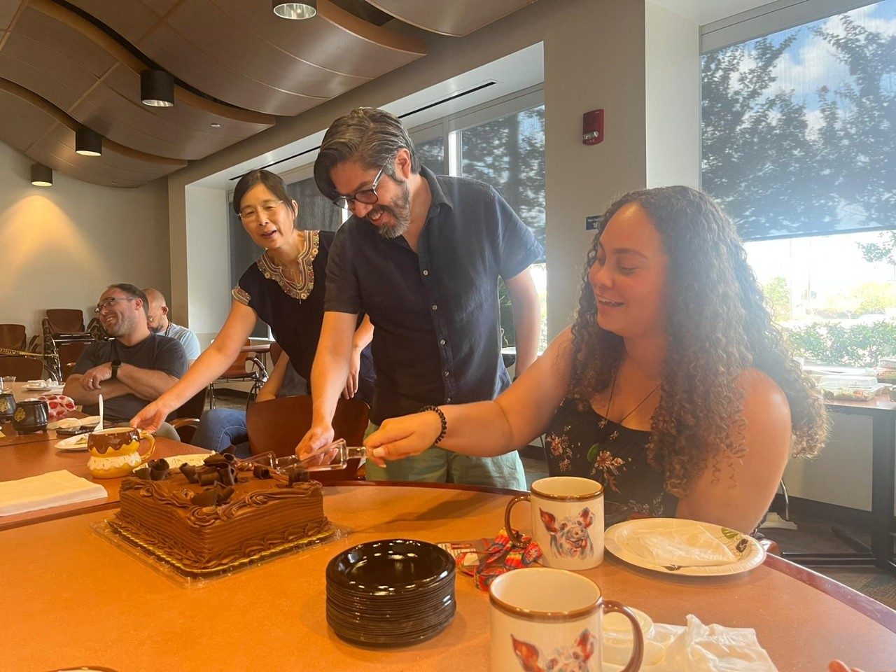 Through people stand by a large chocolate cake, and one of them cuts into the cake