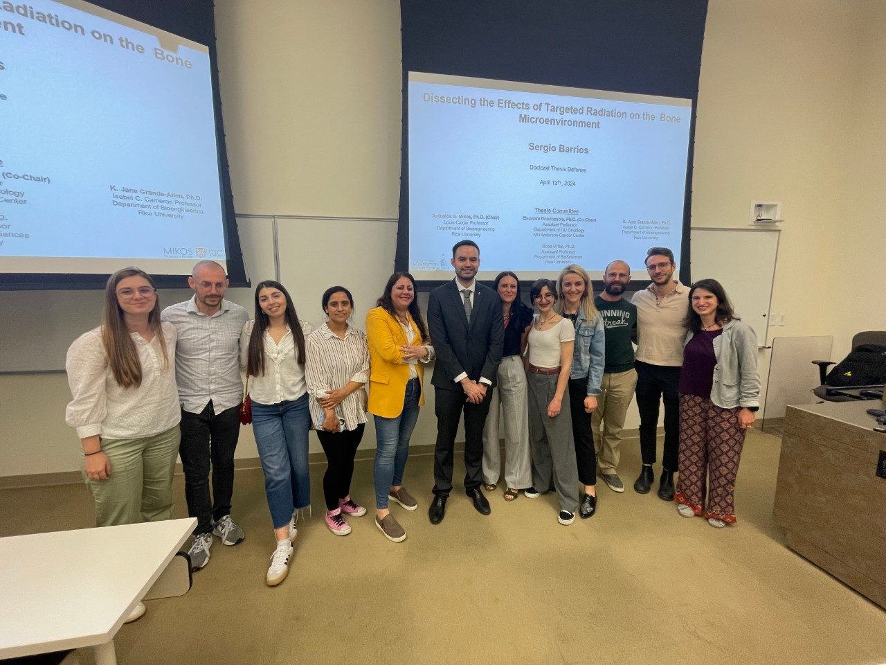 Twelve people posed in one row in front of two screens with PowerPoint presentation defense slides behind them