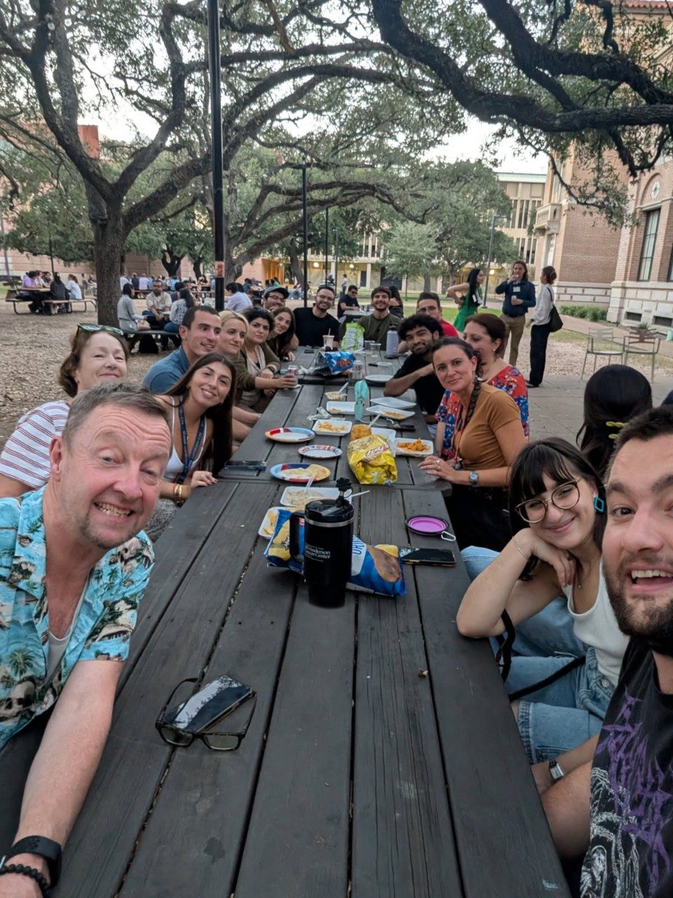 Selfie of a large group of people seated around several picnic tables arranged in a long row