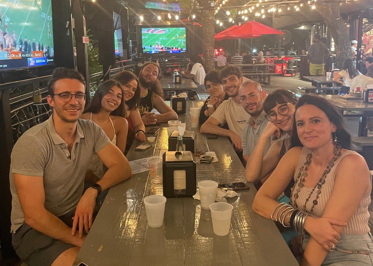 Group of people seated at a long table with water glasses and TVs in the background