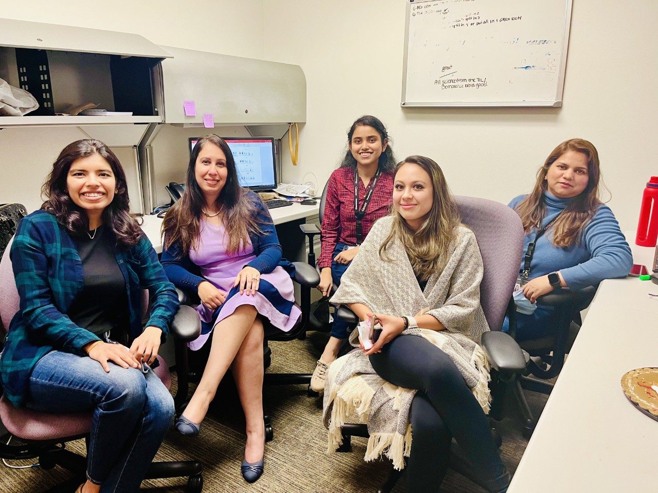 Five women sit in computer chairs in a desk area
