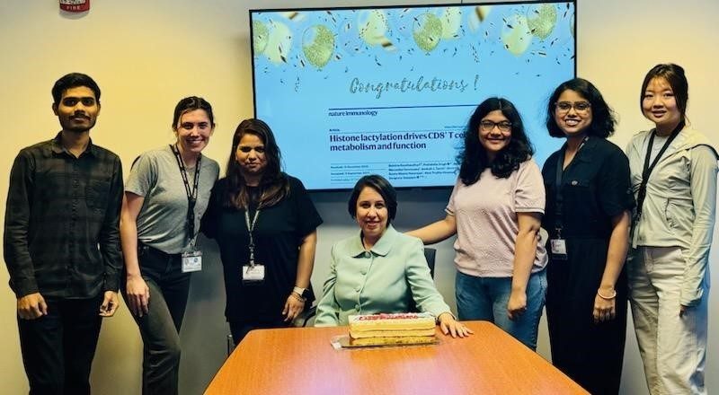 Dr. Goswami is seated at a table with a cake and 6 lab members gathered around her, 3 on each side