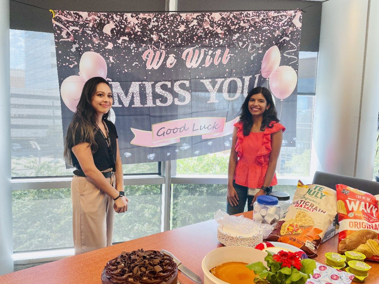 Two women stand in a conference room with a table full of food in front of them and a sign that reads "We Will Miss You" behind them