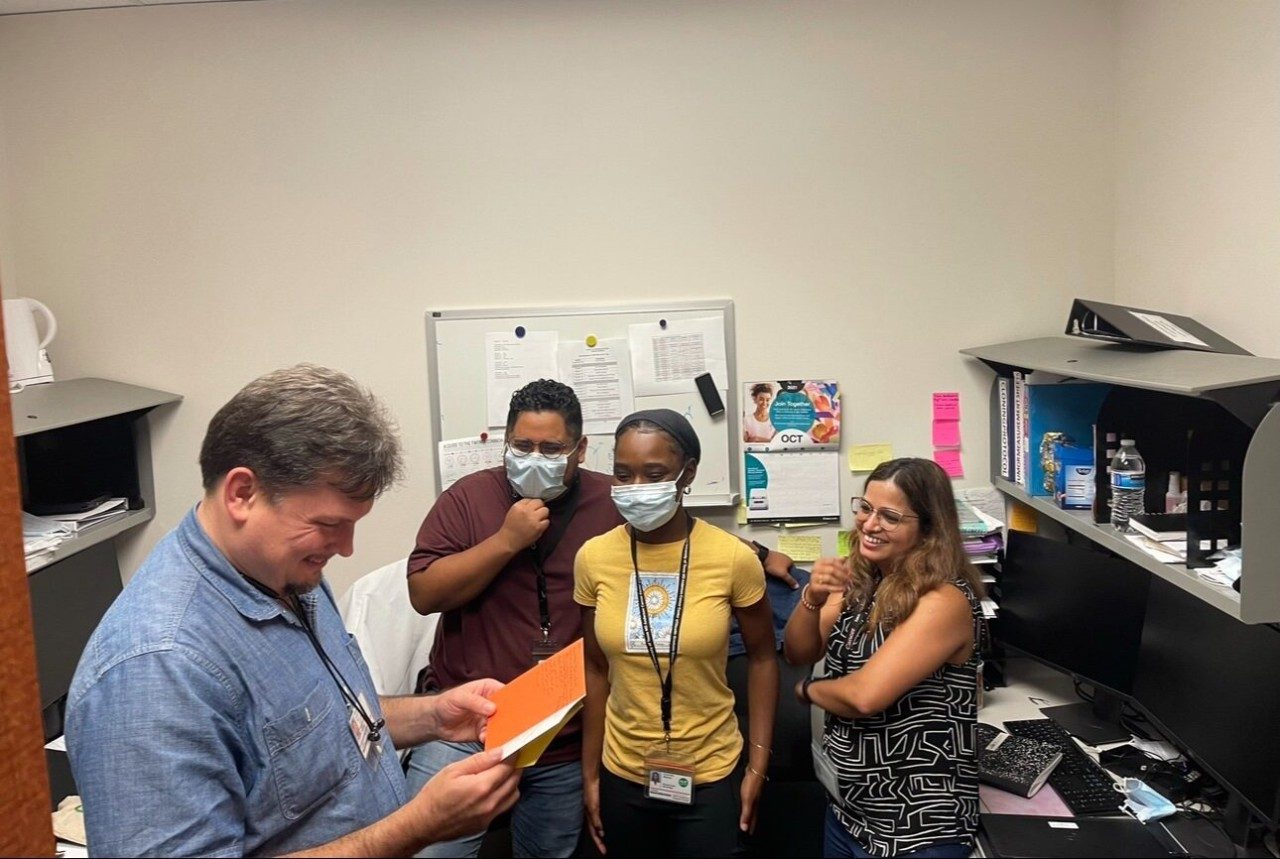 Three people stand and watch as a fourth person reads a card they were given