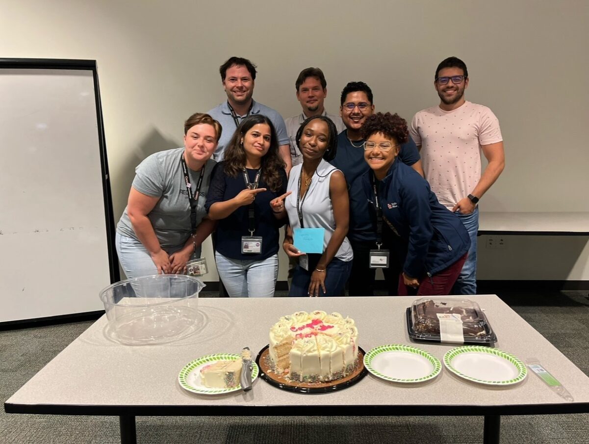 Eight people pose behind a table with a cake on it