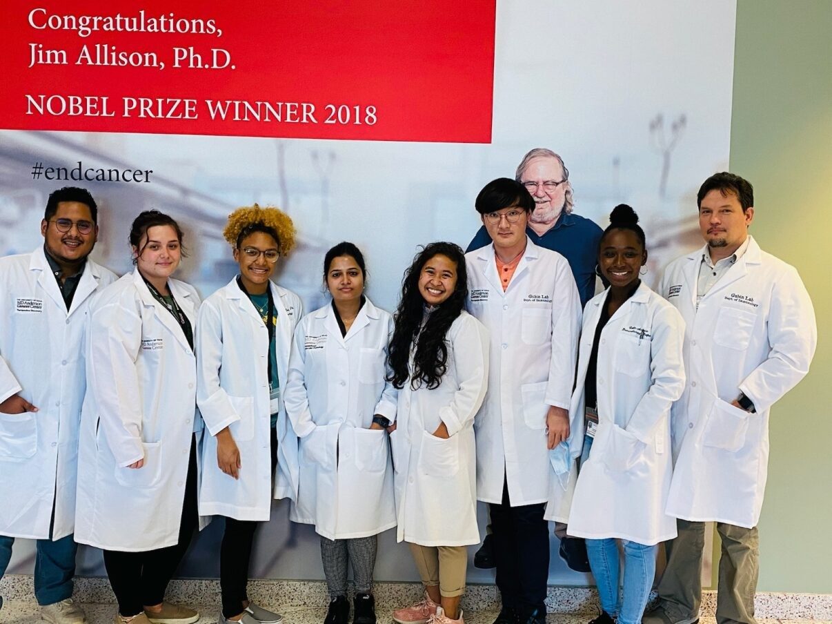 Eight people pose in white lab coats in front of a Jim Allison Nobel Prize Winner sign