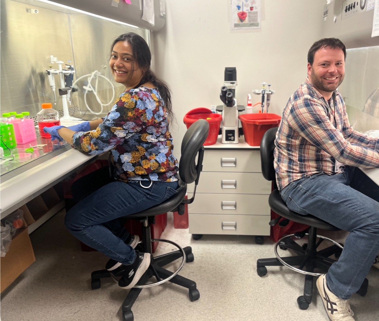 Two researchers pose and smile while working in hoods across from one another