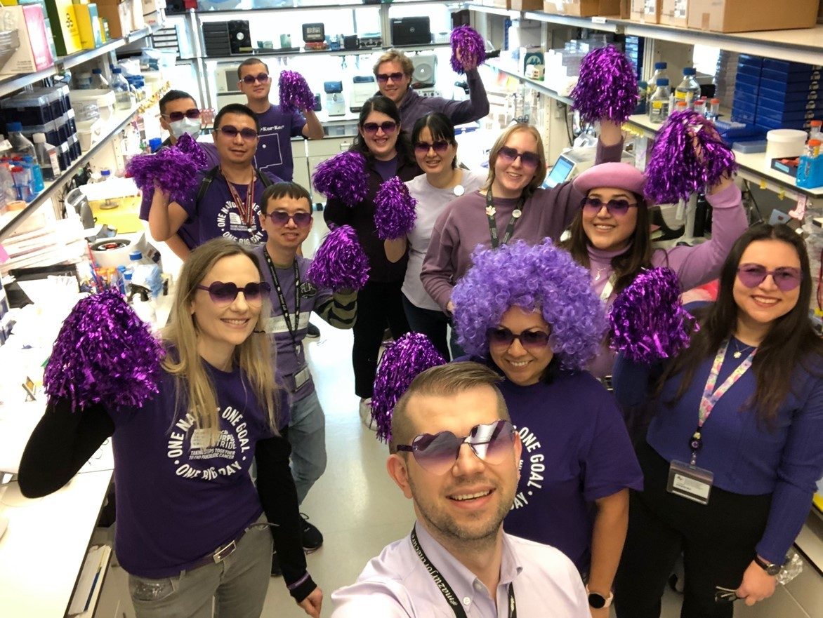 Mazur Lab members standing in a wet lab wearing purple shirts and sunglasses and holding up purple pom-poms