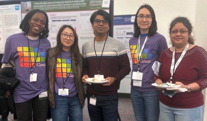 Photo of five Pan Lab members wearing name badges around their necks, posing in front of scientific posters