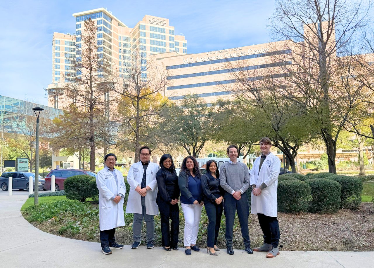 Seven people pose outside, three in white lab coats, in front of green space with the MD Anderson R. Lee Clark Clinic building in the background