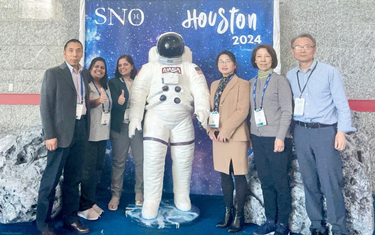 Six lab group members posed in front of SNO Houston 2024 photo backdrop, with an astronaut suit in the center and fake moon rocks on either side