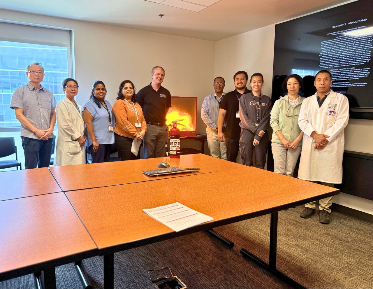 10 people posed standing around a conference room table, with a screen in the back displaying a fire