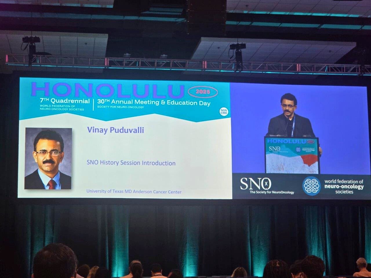 Dr. Puduvalli behind a podium at SNO 2025 on a screen next to his slide for the SNO History Session Introduction