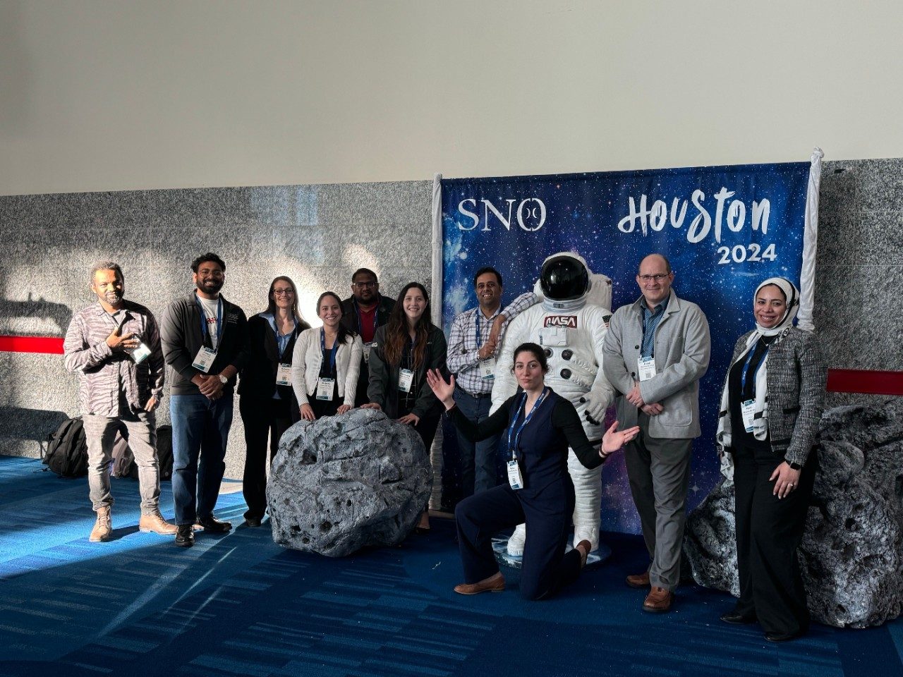 Lab members gathered around a SNO Houston 2024 photo backdrop that includes an astronaut