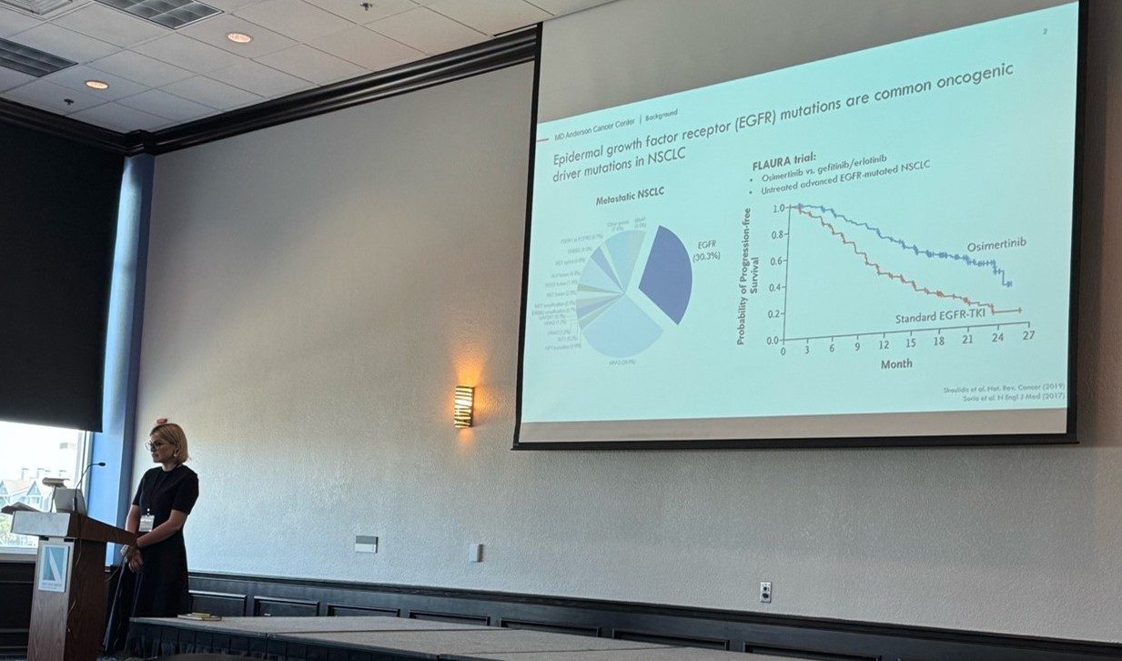 A woman stands behind a podium with a slide on the projector screen