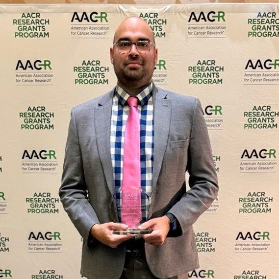 Dr. Reuben in front of an AACR branded wall in a grey suit with a plaid shirt and pink tie