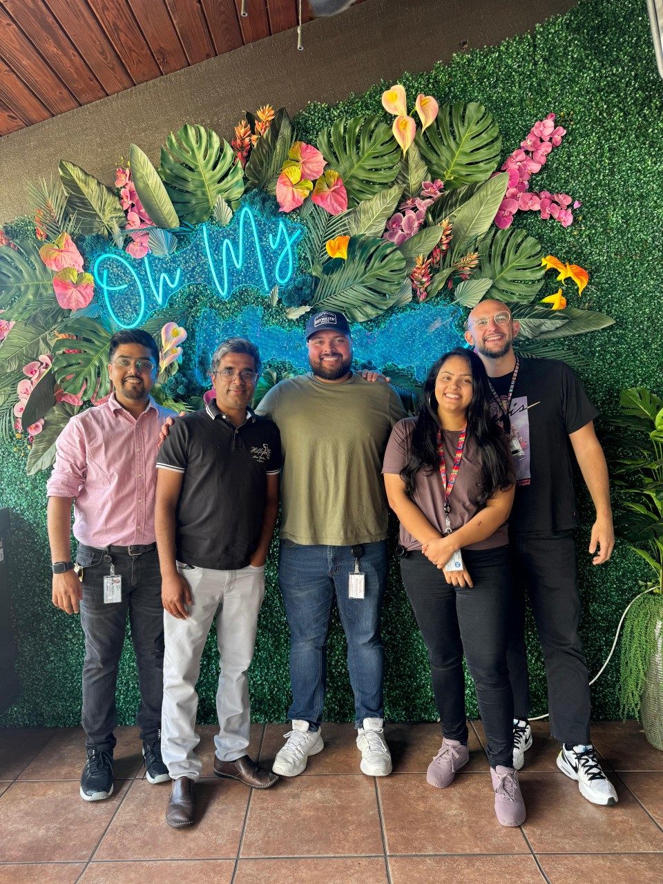 Five people standing in front of a flower and greenery photo backdrop wall that says "Oh My" in blue cursive
