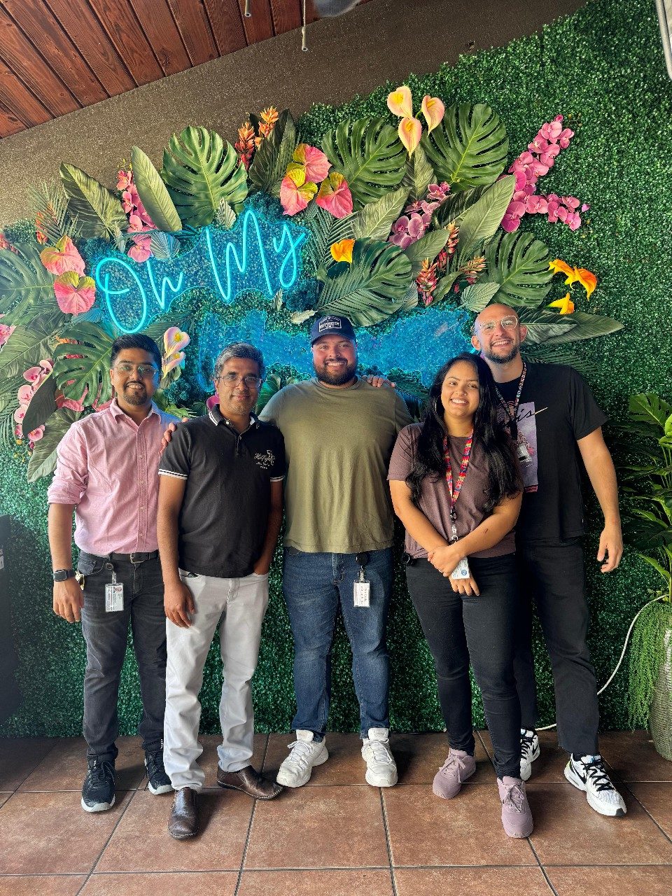 Five people standing in front of a flower and greenery photo backdrop wall that says "Oh My" in blue cursive